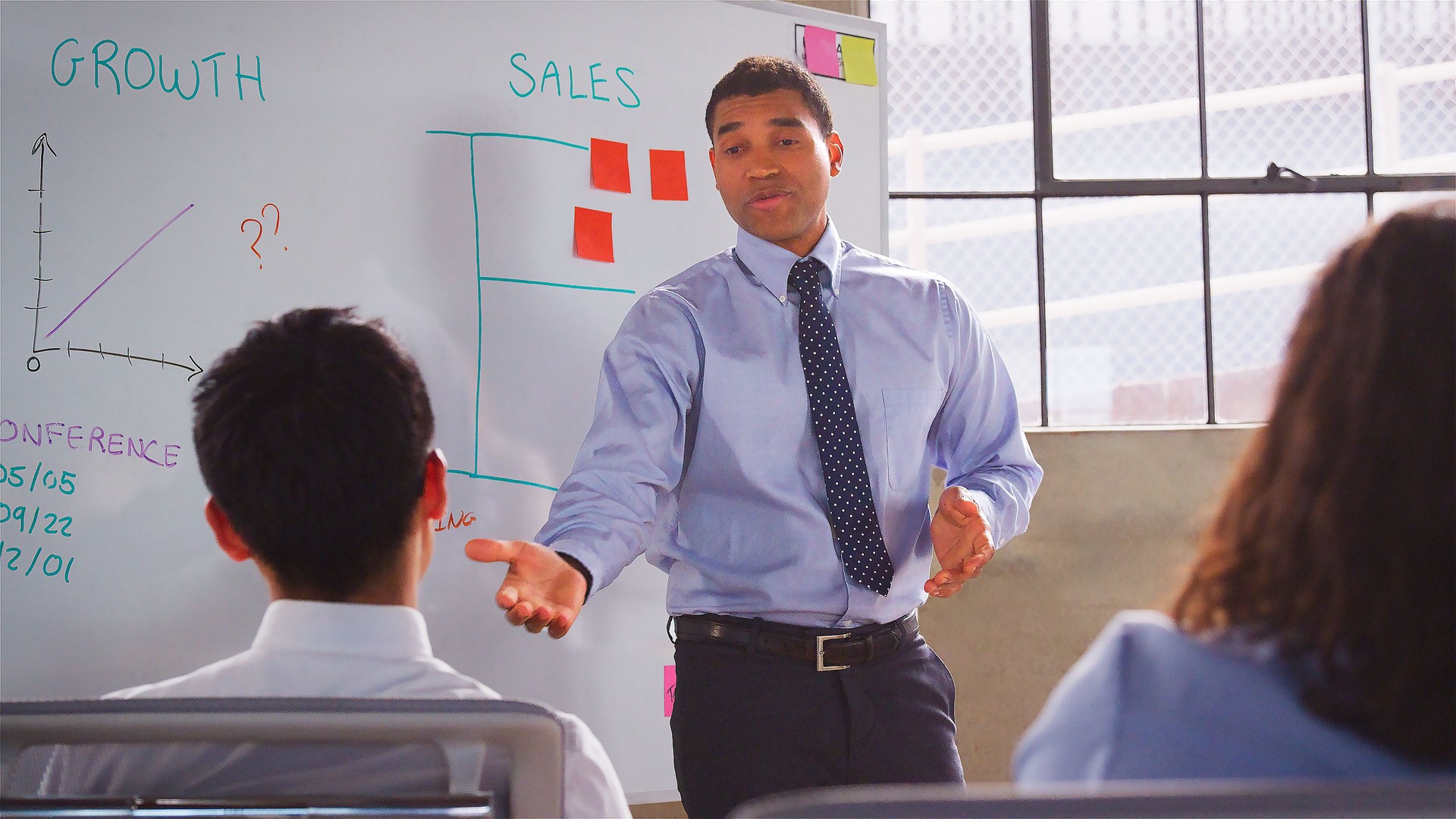 Businessman Standing At Whiteboard Giving Sales Presentation To Multi-Cultural Business Team